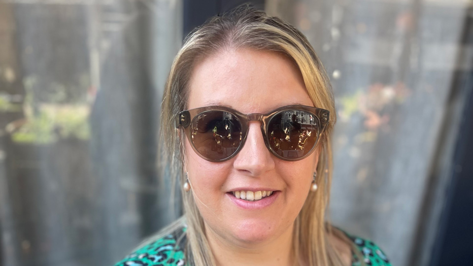 In a green leopard print dress and large brown sunglasses, Leanne smiles and holds a glass with tropical fruit arranged inside
