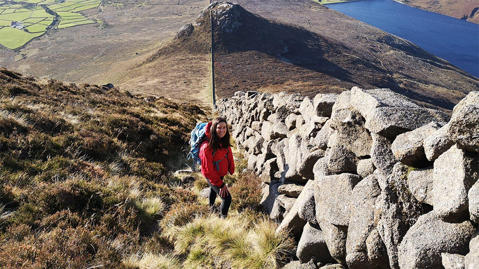 A woman wearing a red jacket and carrying a large hiking backpack climbs a grassy, rocky hillside beside a long stone wall. She smiles toward the camera, with expansive patchwork fields, a mountain ridge, and a deep blue lake stretching out in the background.