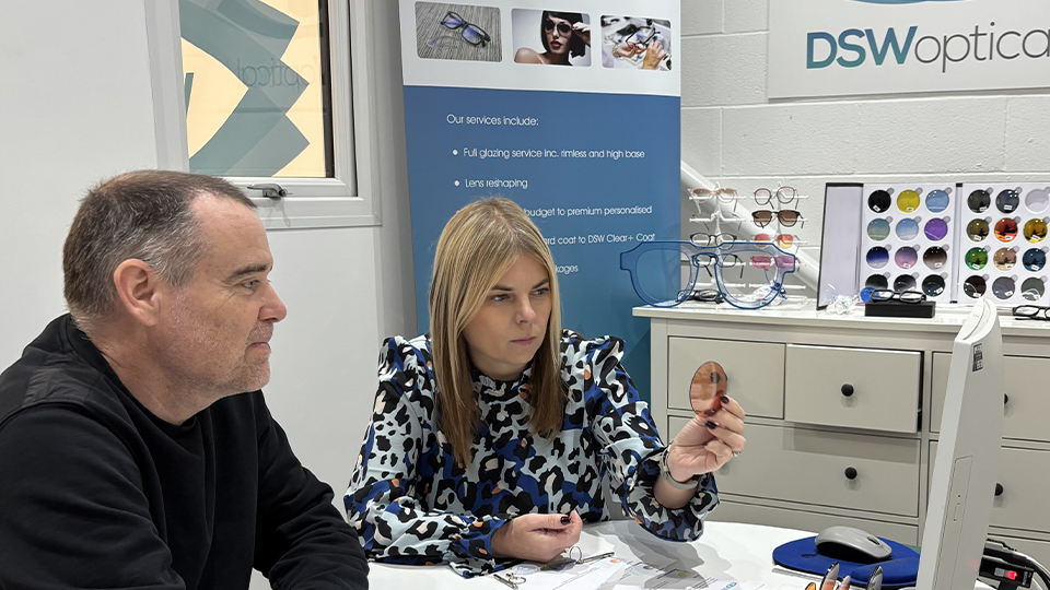 David checks information on a sheet of paper while Mariola holds up a warm-tinted lens. On the table is an array of colourful lenses, Transitions demo kit, and in the background is a banner and sign for DSW Optical and displays of eyewear