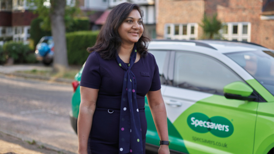 A smiling woman stands on a residential street wearing a navy dress and a matching polka-dot scarf. Behind her is a green and white Specsavers car parked along the curb, with houses and trees in the background