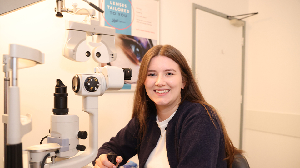 A smiling woman sits in an eye-exam room beside a slit-lamp microscope, holding one of the instrument controls. She wears a dark cardigan and white top, and the clinical room has posters and equipment in the background 