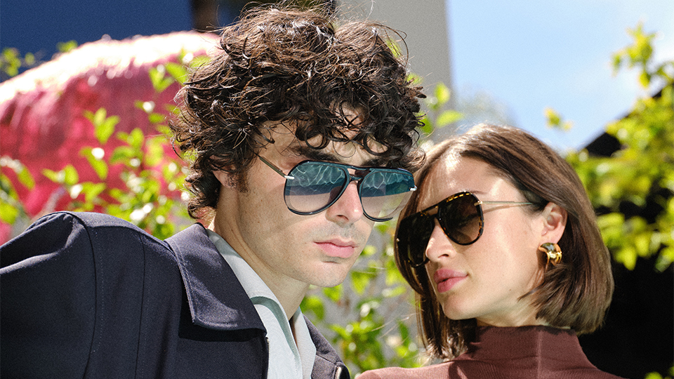 A man in a white shirt and navy jacket with oversized pilot-style sunglasses. The lenses reflect the perfect blue ripples of a pool in the sun. To his right, a woman with short brown hair wears a chestnut brown tight fitting top and pilot sunglasses in brown