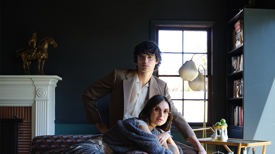 A woman lays on a sofa, wearing a flowing metallic silk skirt and sparkly top, with a soft wool jumper casually around her shoulders and gold glasses with a warm tinted lens. She leans against a man who wears a medium brown suit, an off-white shirt and deep brown loafers. He also wears large squared spectacles with thin metal frames. The room is a dark petrol blue, modern, with a wood-and-glass drinks trolley by a large bookcase