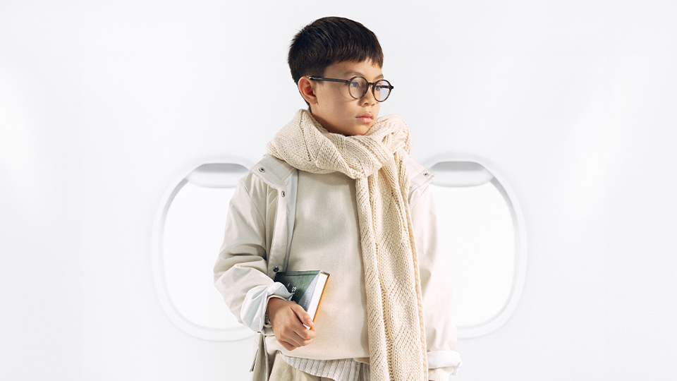 A young boy is dressed in layers of beige clothes and a cream knitted scarf down to his knees for a swaddled but casual appearance. Tucked into one arm is a book. He wears round spectacles. Behind him are two plane windows.  