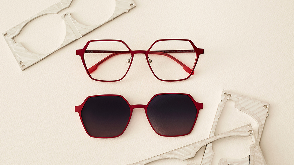 Against a pale cream background is a spectacle frame and a clip-on sunglasses lens made of a thin matte recycled aluminum. The frames are a polygon shape in a vibrant raspberry red. Positioned around it are offcuts of material sheets, presumably from which the frame was cut