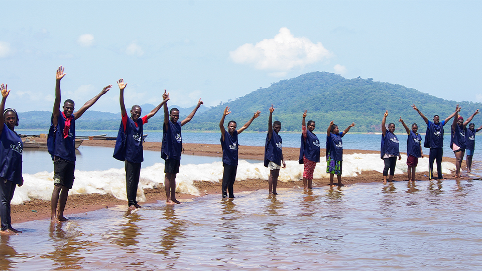 A group of people who are part of the Sea2see plastic recycling initiative stand in a line along a shoreline waving at the camera