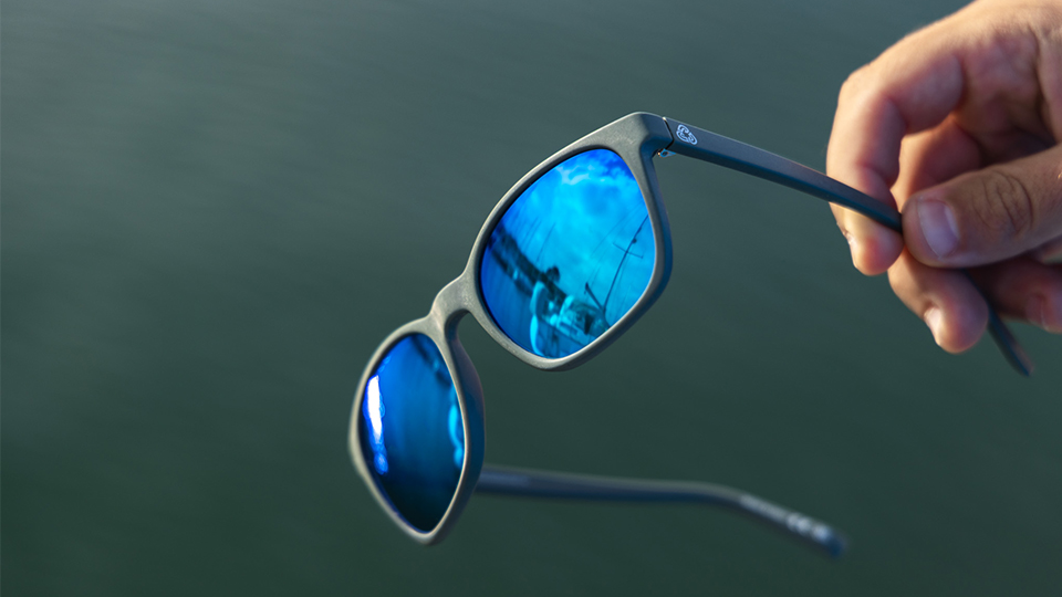 A pair of grey sunglasses with blue mirrored lenses are held out above the blue-green of the ocean