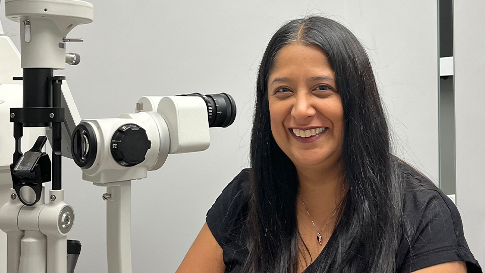 Kavita is wearing a black top and smiling with her arms folded on a table in a consulting room