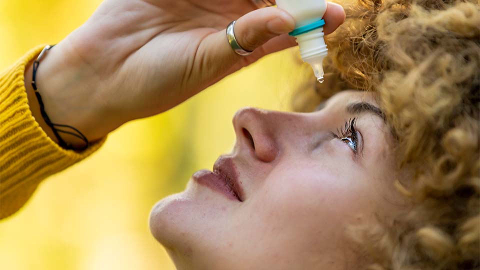 Close up of a woman wearing a mustard cardigan and using eye drops. She has curly hair   