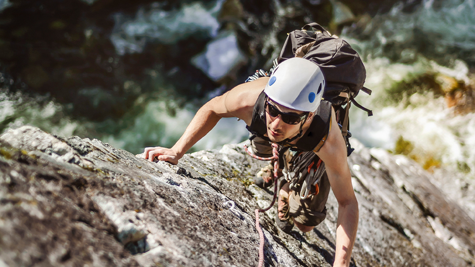 A climber, wearing a protective helmet and Hoya Sportive sunglasses, scales a rockface