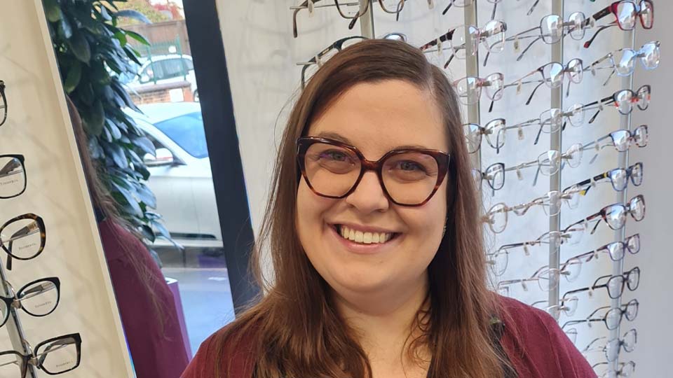 Emily is seated behind a desk in practice with a computer to her right and a mirror reflecting the rows of spectacles on her left 