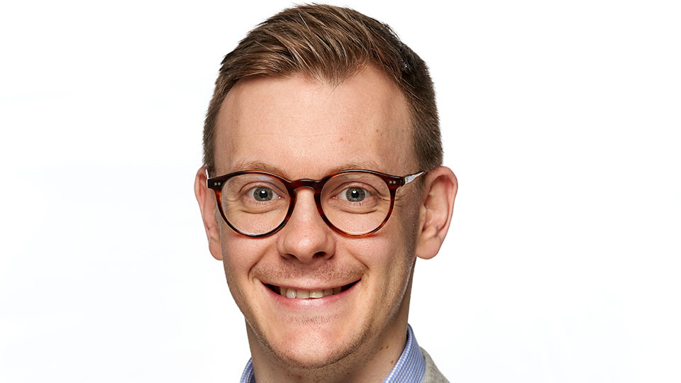 Dr Grant Robinson is smiling at the camera whilst wearing a blue shirt, grey suit jacket and tortoiseshell glasses  