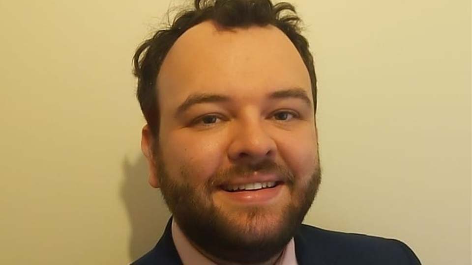 James Brawn is smiling at the camera in front of a yellow wall 