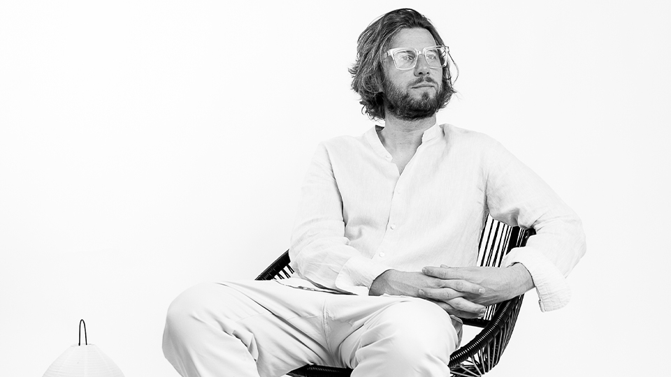 Marco, dressed in a light shirt and chinos, relaxes in a wicker chair, wearing large square spectacles with thick rims. On the floor beside his chair is a brand catalogue