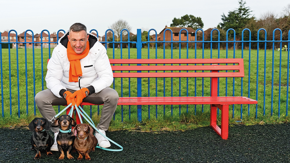 Neil Retallic sits on a park bench smiling. He holds the leads of three dogs that sit in front of him 