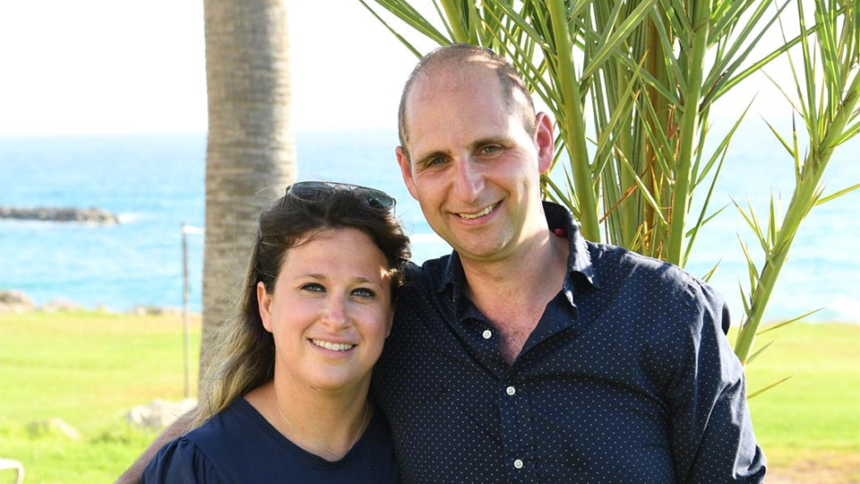 Malya and Adam stand together in a beautiful coastal landscape, palm trees and light blue sea behind