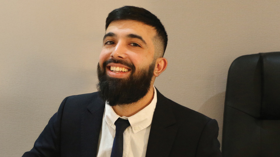 Ali Mahmood is wearing a navy blue suit and sitting at a desk smiling