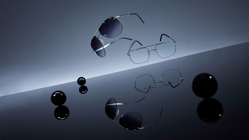 A pair of aviator sunglasses and spectacles against a gradient grey and black background and reflective surface