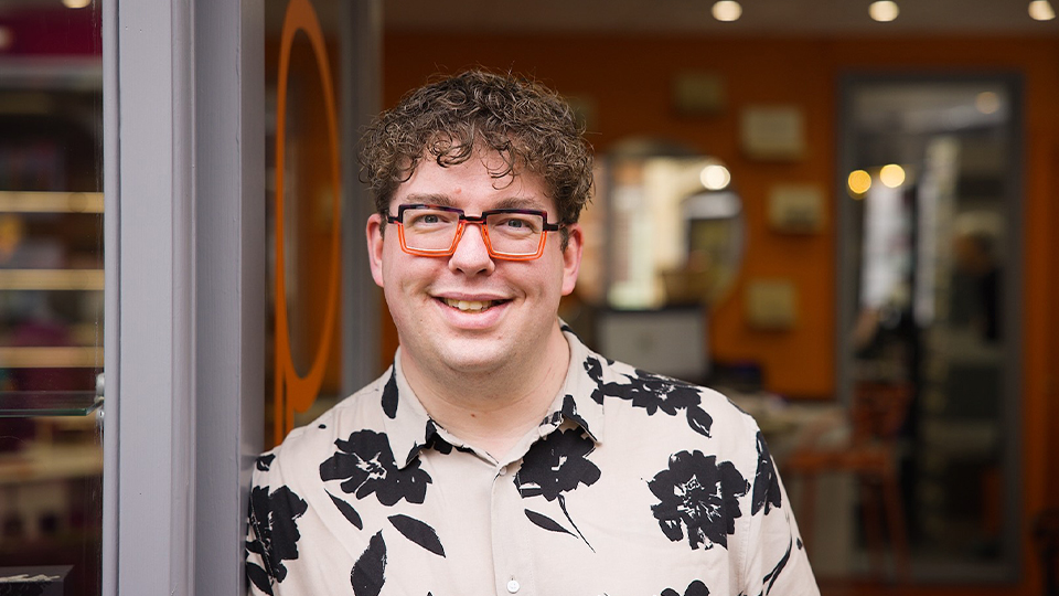 Chris, wearing a floral black and white top and frames in black and orange, stands in the practice doorway 