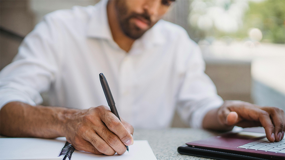 A man makes notes in a notebook with a laptop open beside him