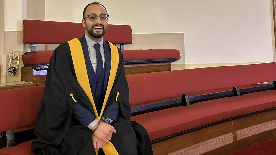 Jag is wearing a graduation gown and is sat in front of a College of Optometrists sign 
