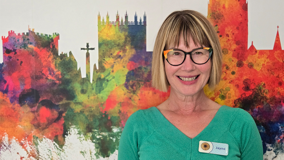Jayne wears a green top with a name badge and black glasses with orange and yellow geometric details. She stands in front of a wall with a watercolour skyline