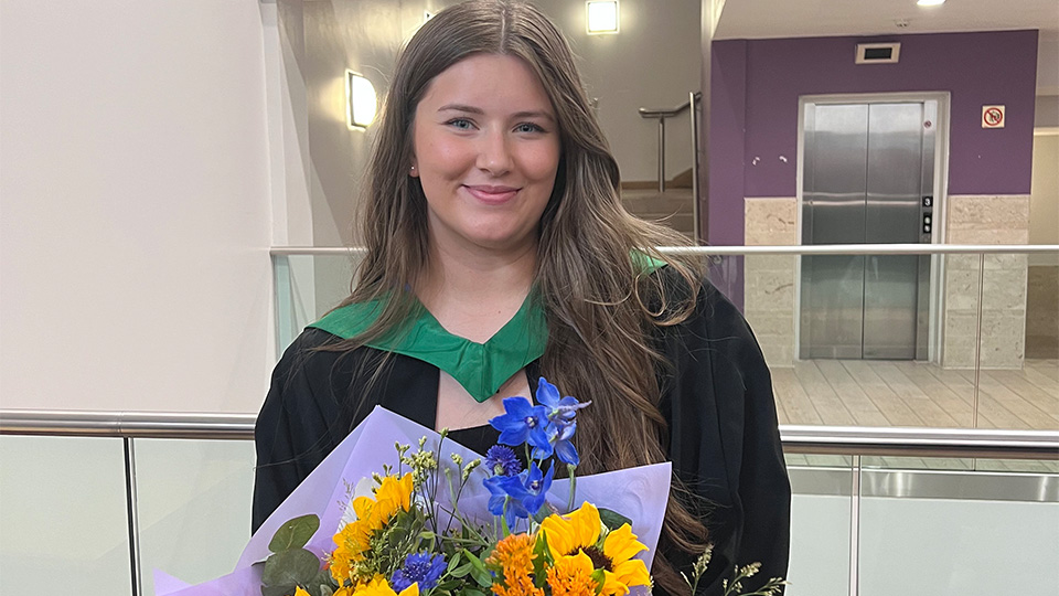 Eliza is in graduation robes and is holding a bunch of sunflowers