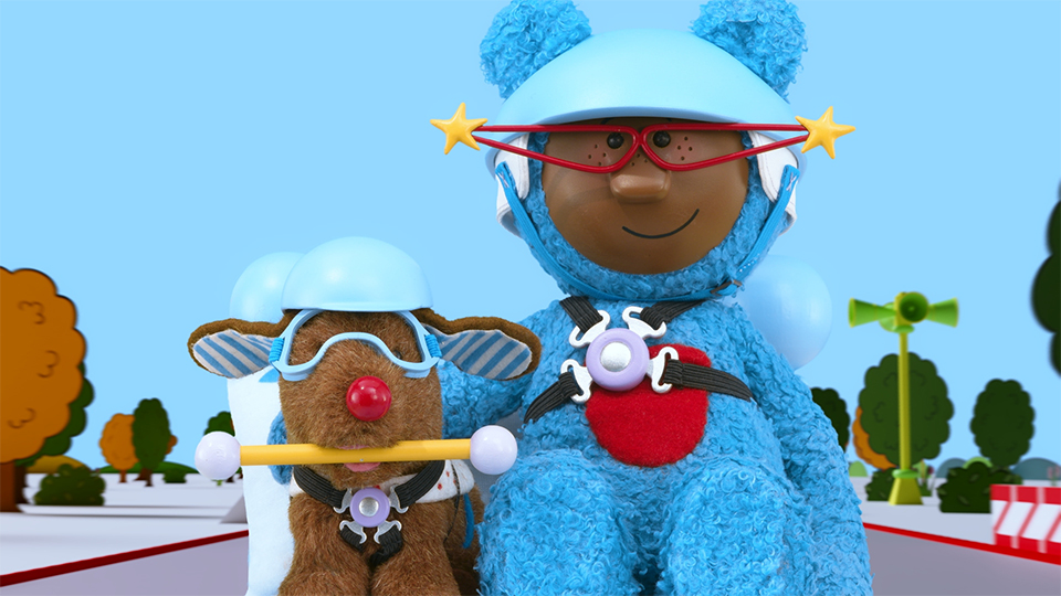Pockets the bear on the right wears a helmet and is strapped in to a vehicle. On her left is Yapette her guide dog