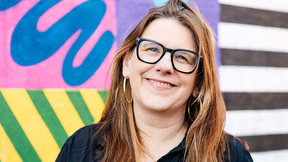 Standing in front of a colourful patterned background, Rebecca wears large squared glasses with a thick black frame. She has brown hair and large gold hoop earrings, a navy top and jeans.