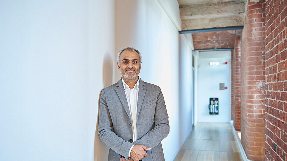 Dharmesh has his hands crossed and stands in front of a wall wearing a grey suit