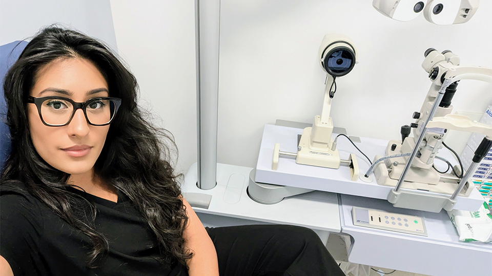 Aminah has black hair and is looking towards the camera whilst sat at her desk in a consulting room