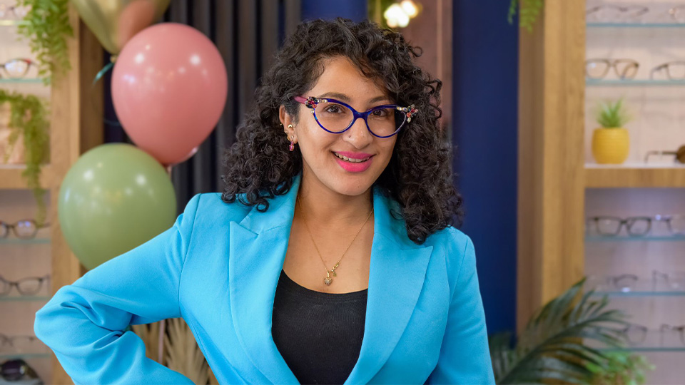 Rizwana poses in a colourful practice with pastel balloons on the wall. She wears a light blue blazer and navy cat’s eye style frames, with gems in the upturned corners, and pink temples