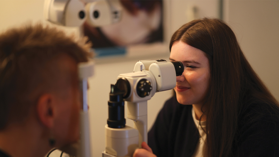 Young female optometrist smiling as she looks through a slit lamp microscope to examine a male patient’s eyes in a softly lit clinic room, with a blurred owl picture on the wall in the background
