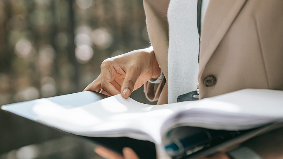 Camera is focused on the torso of a person in a brown suit jacket holds open a binder of documents and leafs through them.
