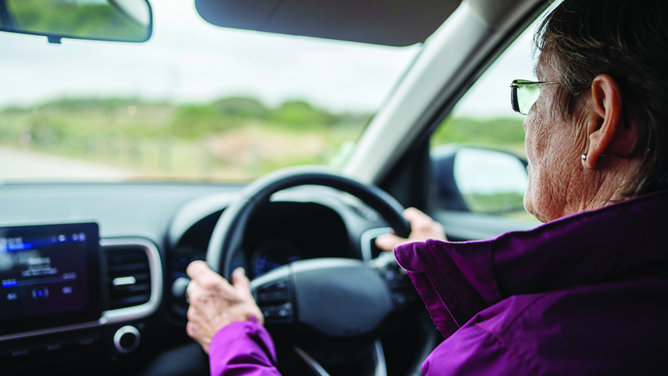 The hand of an older driver rests on the steering wheel while driving. She wears a pink coat and has glasses.