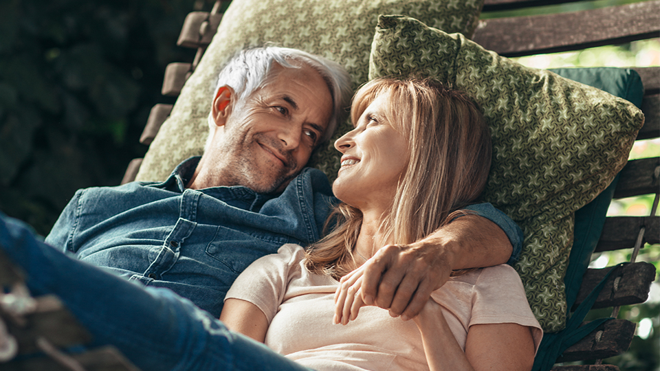 A mature couple lying on a wooden slatted hammock, both in casual clothes and smiling at each other