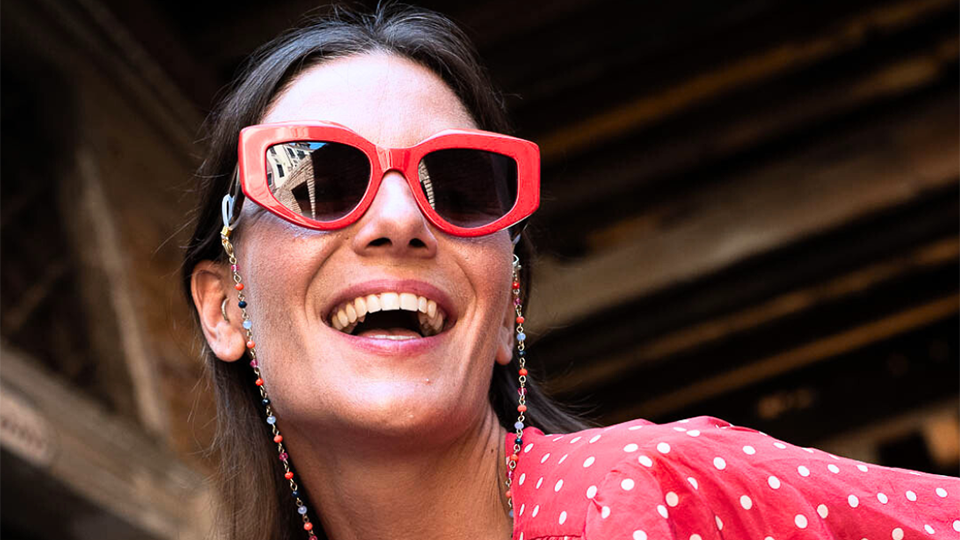 A model looks summary and happy wearing a red top, red rectangle sunglasses and a rainbow beaded glasses chain