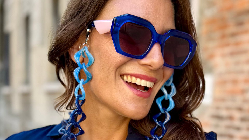 A model laughs happily, she is wearing oversized pink and navy sunglasses with a chunky glasses chain in a gradient from light blue to navy