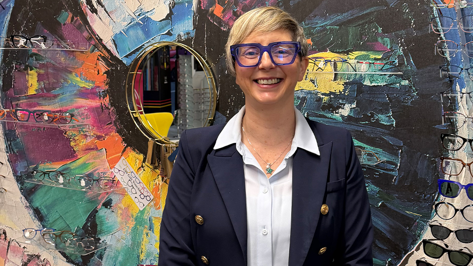 Lindsey Jones smiling whilst stood in front of a large colourful mural inside her optometry practice