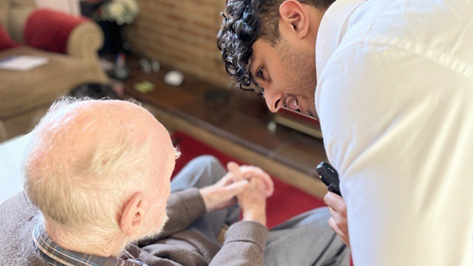 A healthcare professional leans over to examine an elderly man seated in an armchair in a living room, holding a handheld diagnostic device close to the patient’s eye. The room has patterned wallpaper, a wooden cabinet, and soft furnishings, creating a warm, homely setting