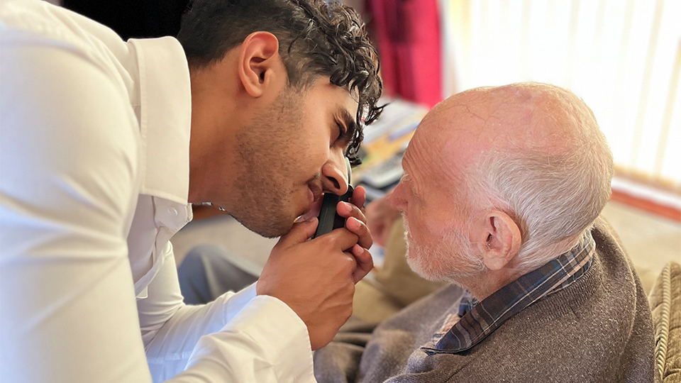 A healthcare professional leans in to examine an elderly man’s eye using a handheld ophthalmoscope as the man sits in an armchair at home, with soft daylight and living room furnishings in the background