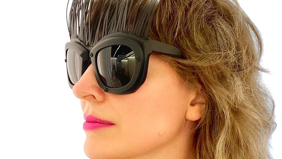 Kayla looks off to the left of the frame wearing a pair of oversized black sunglasses in a mask-like style. From the browline emerge tens of metal antenna that end in little rings. The metal fades from black at the browline to silver. Kayla wears a hot pink lipstick and a neon patterned top
