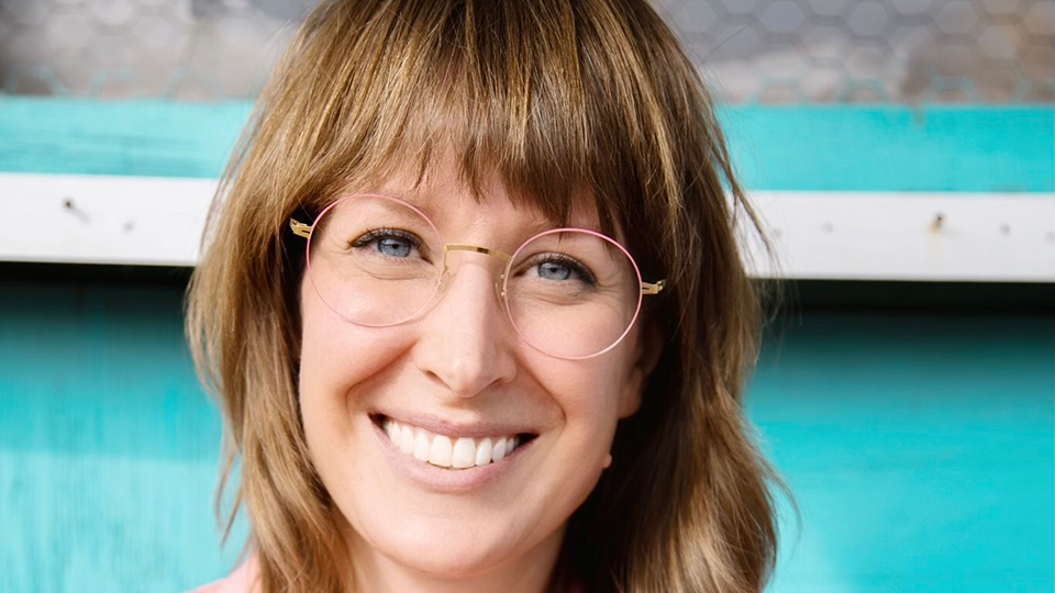 Kayla has brown hair cut in a layered style just past her shoulders. She wears a round pair of spectacles with metal temples and a very thin light pink rim. She wears a pink work shirt with a patch of a stylised skull
