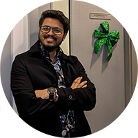 Guru is smiling with his arms folded whilst leaning against a wall