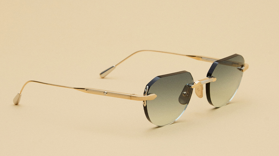 A pair of rimless spectacles on a beige background. The frames are a gold metal with bevelled green ombre lenses
