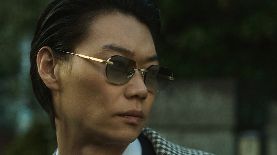 Japanese actor, Show Kasamatsu, models a pair of metal spectacles with rimless green lenses. He wears a white shirt with sharp collar, a yellow and green patterned tie, and dark suit jacket, over the top he wears a green-grey houndstooth coat.  