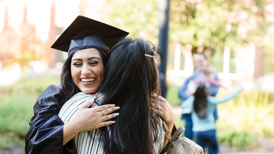 A mother embraces her daughter, who is wearing a graduation gown and cap  
