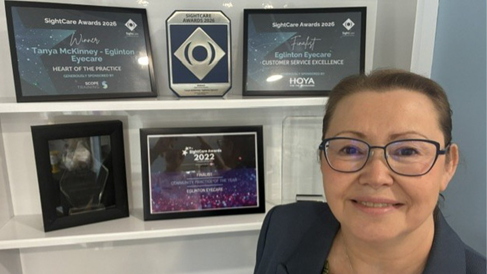 Tanya is standing in front of shelving displaying six award recognitions, including a certificate and a trophy for Heart of the Practice. Tanya has her brown hair tied back, wearing a green plaid top with a navy blazer