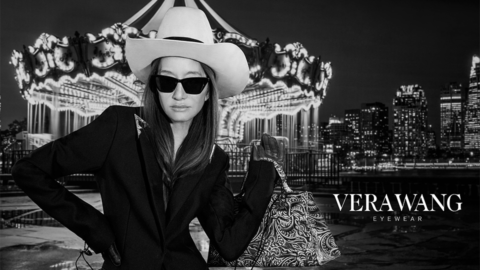 Vera is photographed in black and white wearing a tailored black suit jacket, angular sunglasses, and a pale cowboy hat. One hand is on her waist and the other holds a paisley handbag. Behind her is a carousel, lit up against the night sky, as well as a sparkling cityscape