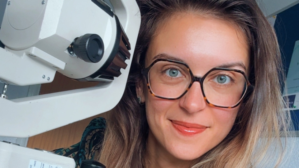 Desislava leans around the side of a slit lamp to take a selfie. She has hexagonal glasses frames, a colourful paisley blouse and bright pink nails